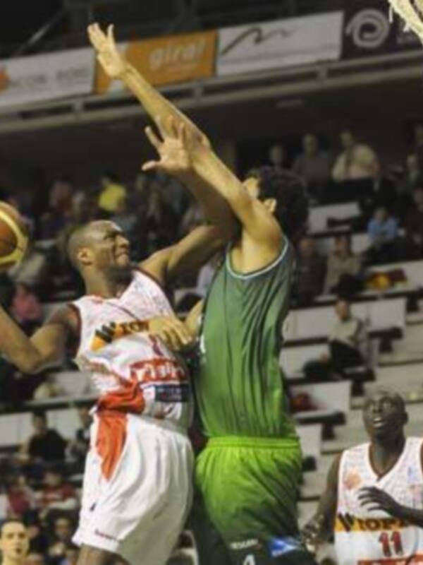 Un jugador de baloncesto intenta anotar mientras otro intenta bloquear el tiro. El jugador blanco lleva una camiseta con el nombre 'Villena' en la espalda, mientras que el jugador verde lleva una camiseta con el nombre 'Villena' en la espalda.