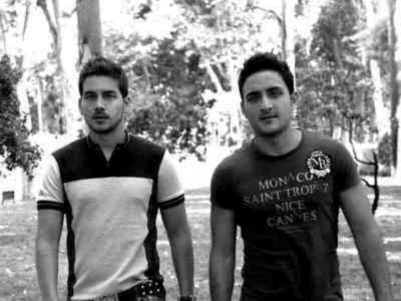 Dos jóvenes posan en un parque, uno con camiseta de fútbol y otro con una camiseta deportiva. Ambos parecen estar en buen estado de salud, con cabello corto y barba. El fondo muestra árboles y un camino de tierra, sugiriendo un entorno natural.