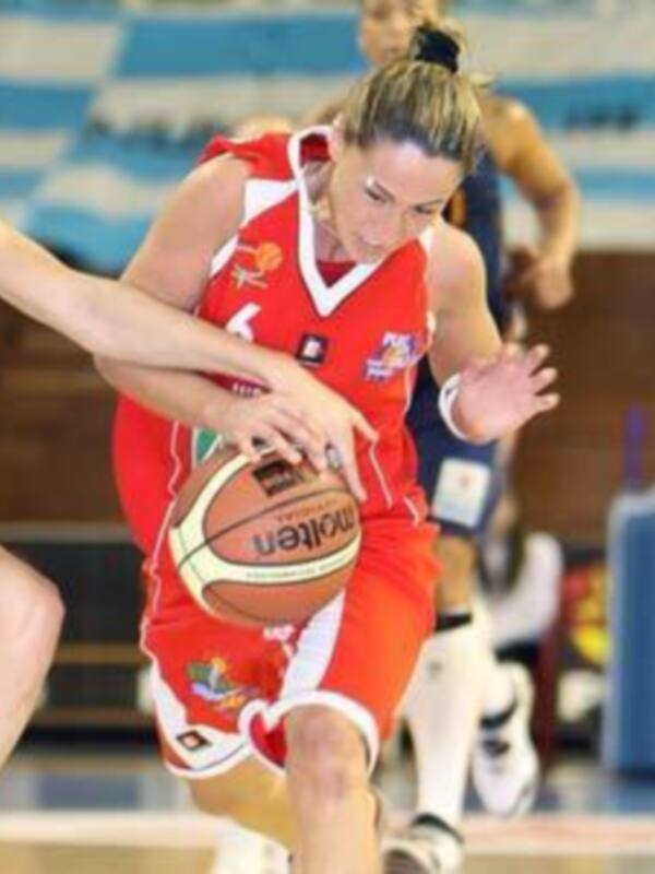 Una jugadora de baloncesto en acción, con una camiseta roja y blanca, controlando el balón mientras otra jugadora intenta desbordarla. El fondo muestra una cancha de baloncesto con líneas y un árbitro en el borde.