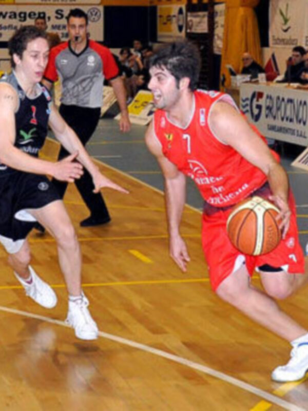 Un jugador de baloncesto en acción, con el balón en la mano, mientras otro jugador lo persigue en un partido de baloncesto en un pabellón deportivo.