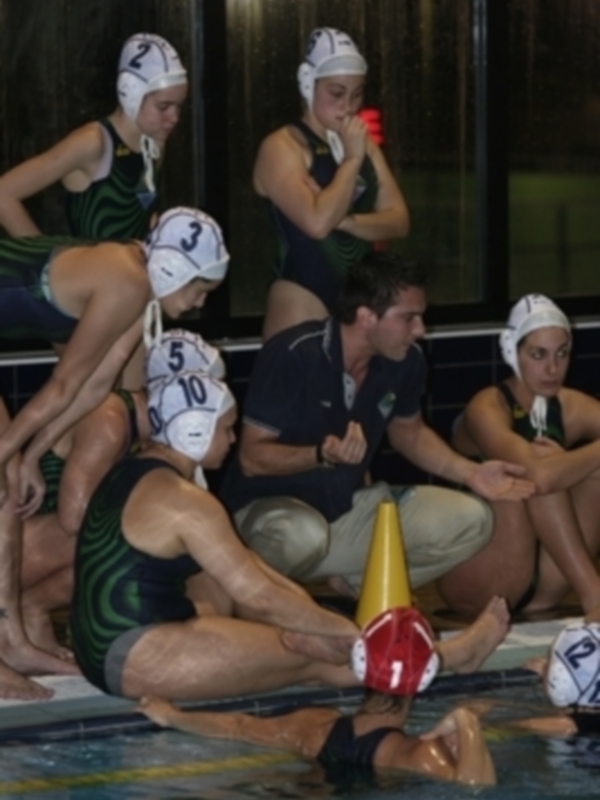 Equipo de waterpolo femenino en acción durante un partido nocturno en una piscina interior, con jugadores vestidos con uniformes de color verde y negro, y un entrenador observando desde el borde del agua.