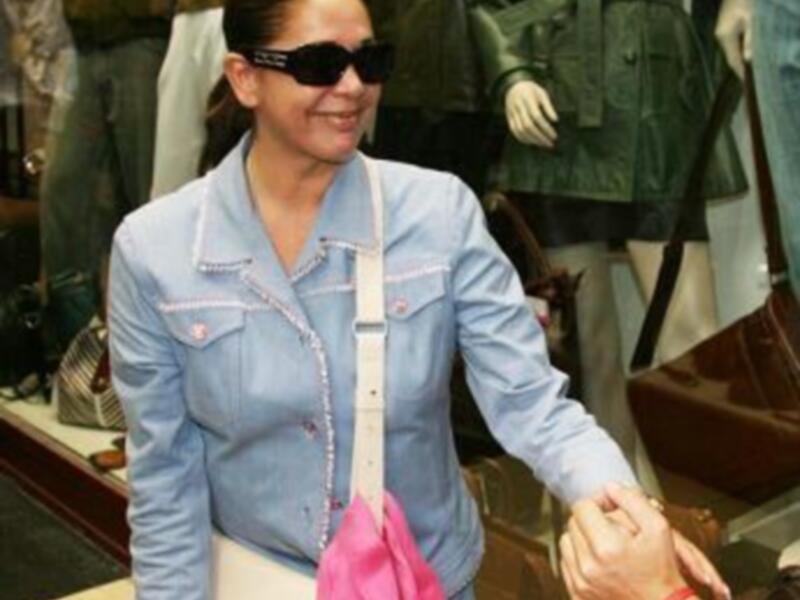 Una mujer con gafas de sol y una chaqueta azul está sosteniendo un bolso rosa mientras camina por un pasillo de tienda.