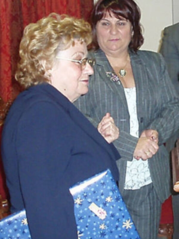 Dos mujeres, una mayor con cabello canoso y otra más joven con cabello oscuro. La mujer mayor sostiene un sobre azul con estrellas y flores, mientras que la joven sonríe. Ambas están en una sala con cortinas rojas y un hombre parcialmente visible detrás.