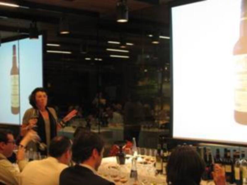 Una mujer en un evento de presentación de productos, con un proyector en el fondo mostrando imágenes de botellas de vino.
