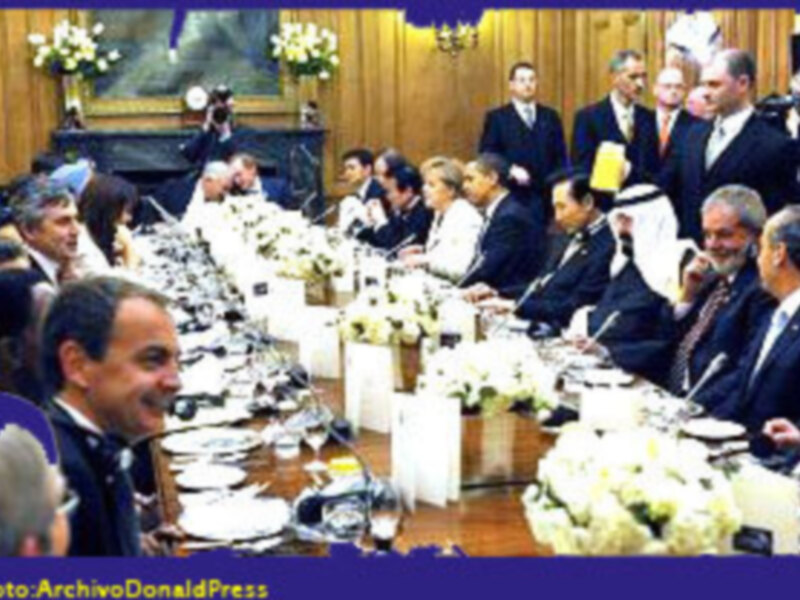 Reunión formal en una mesa larga con participantes en trajes, decoración elegante y flores blancas.