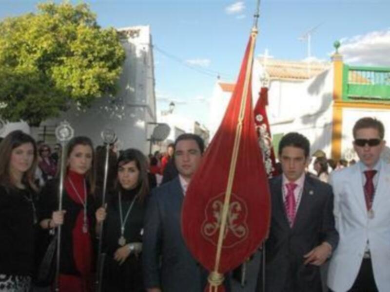 Una procesión religiosa con personas vestidas formalmente, sosteniendo una bandera roja y un círculo dorado. El entorno muestra edificios tradicionales con colores brillantes y un cielo despejado.