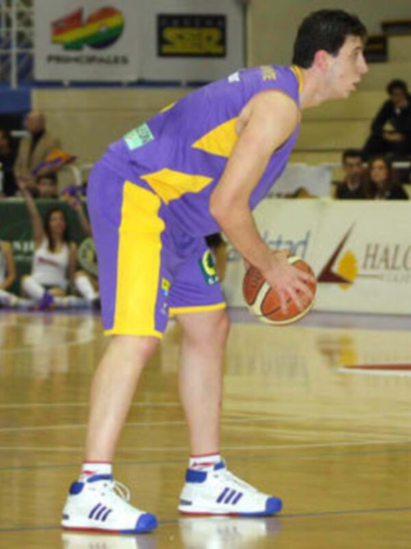 Jugador de baloncesto en acción durante un partido, vestido con uniforme amarillo y lila, controlando el balón en el piso de un estadio deportivo.