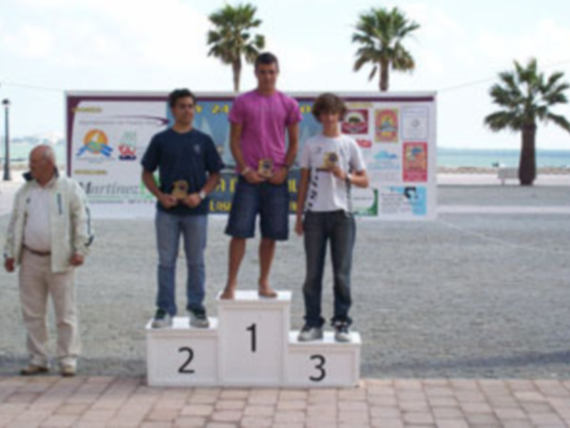 Tres jóvenes en un podio, con un hombre en el fondo, frente a una playa y una señal informativa.