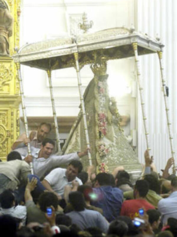 Una multitud se congrega alrededor de una imagen religiosa en un carro, rodeada por flores y luces. La escena parece ser de una celebración o procesión religiosa en un lugar sagrado.