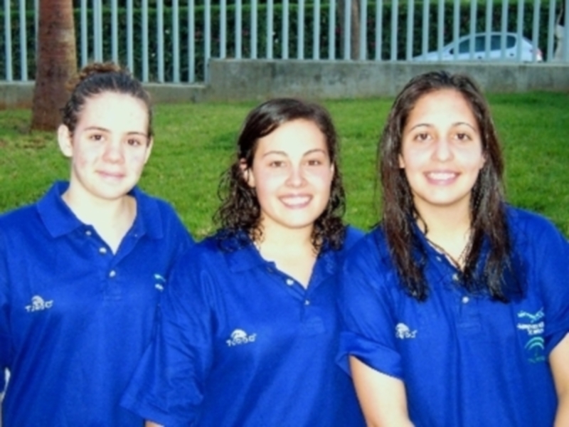 Tres jóvenes en camisetas azules posando para la foto en un parque con un árbol y un edificio en el fondo.