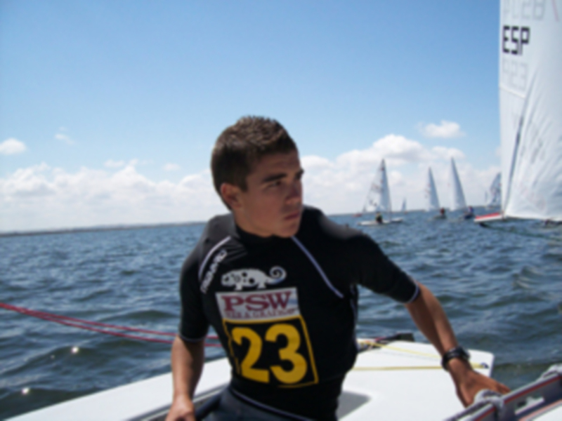 Un joven competidor en una regata de vela, con el número 23 en su chaleco, navegando en el mar bajo un cielo azul.