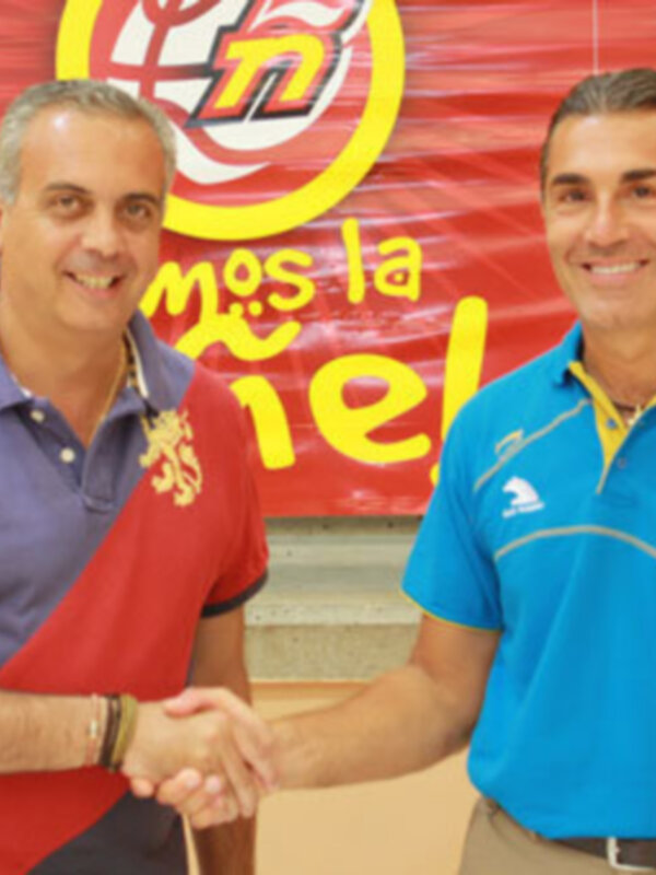 Dos hombres se saludan con una bandera roja y amarilla en el fondo, con el logo de 'Caja Rural' visible.