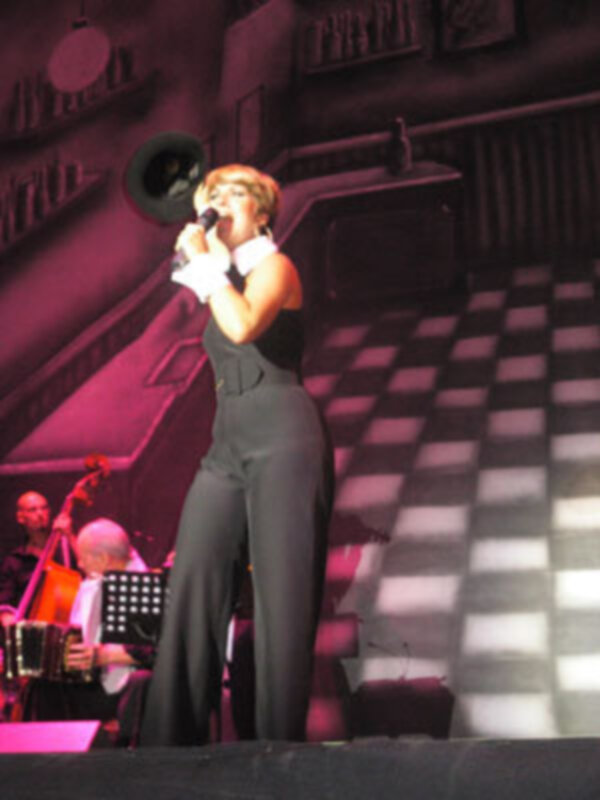 Una mujer con un vestido negro y pantalones largos está cantando en una escena de concierto con un fondo de luces rojas y negras.