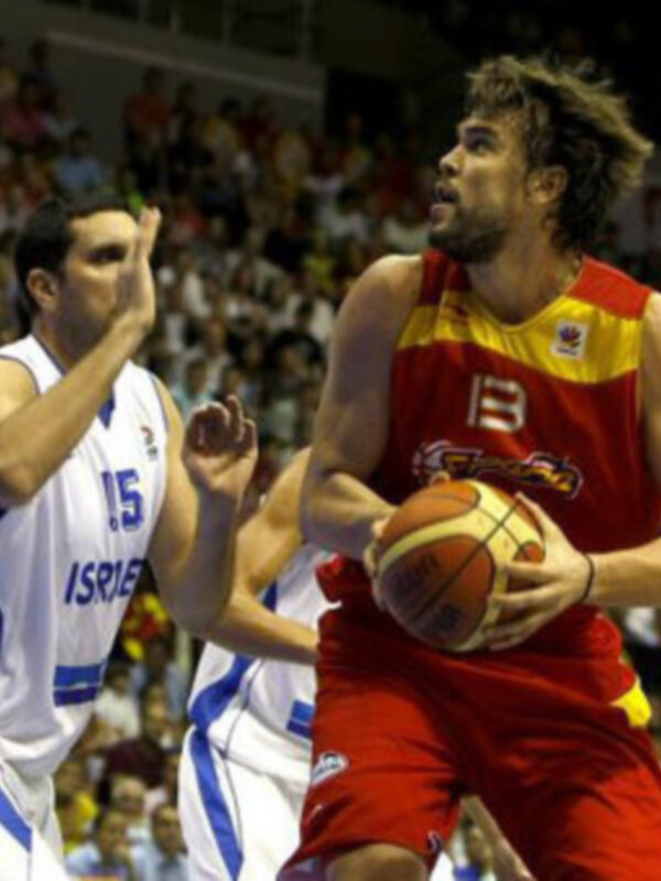 Un jugador de baloncesto en acción, con una camiseta roja y amarilla, controlando el balón mientras un rival en blanco y azul lo persigue.