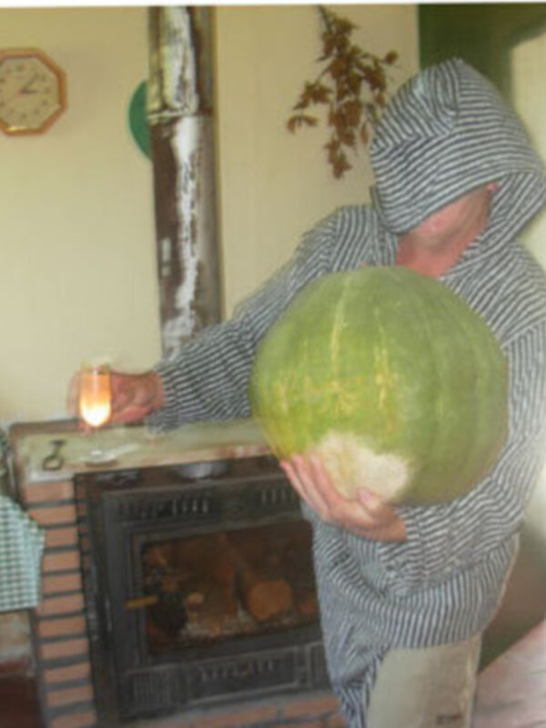 Persona en traje de monje sosteniendo una gran calabaza verde en una habitación con chimenea y reloj en la pared.