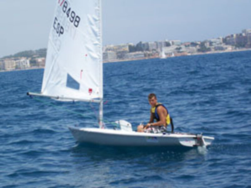 Un hombre navega en un pequeño barco blanco con una vela blanca y azul, navegando en el mar azul profundo. En la orilla se pueden ver edificios y una playa, con el cielo despejado.