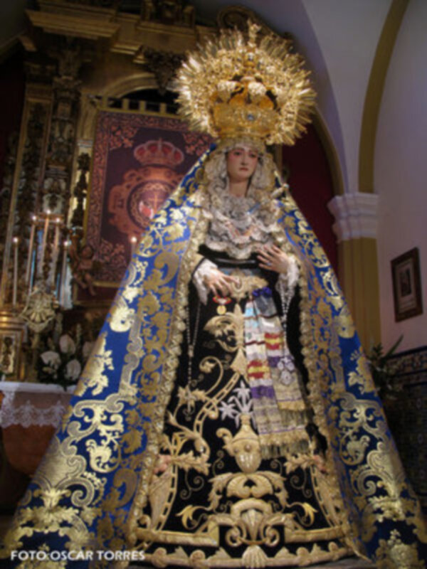 Imagen de una Virgen María vestida con trajes dorados y azules, coronada con un halo de oro. La imagen está en una iglesia con detalles arquitectónicos y una bandera de fondo.