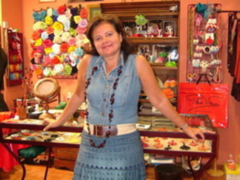 A una mujer sonriendo mientras sostiene un cortejo de flores. Está vestida con un vestido azul y una corona de flores en la cabeza, frente a una vitrina llena de adornos y decoraciones navideñas.