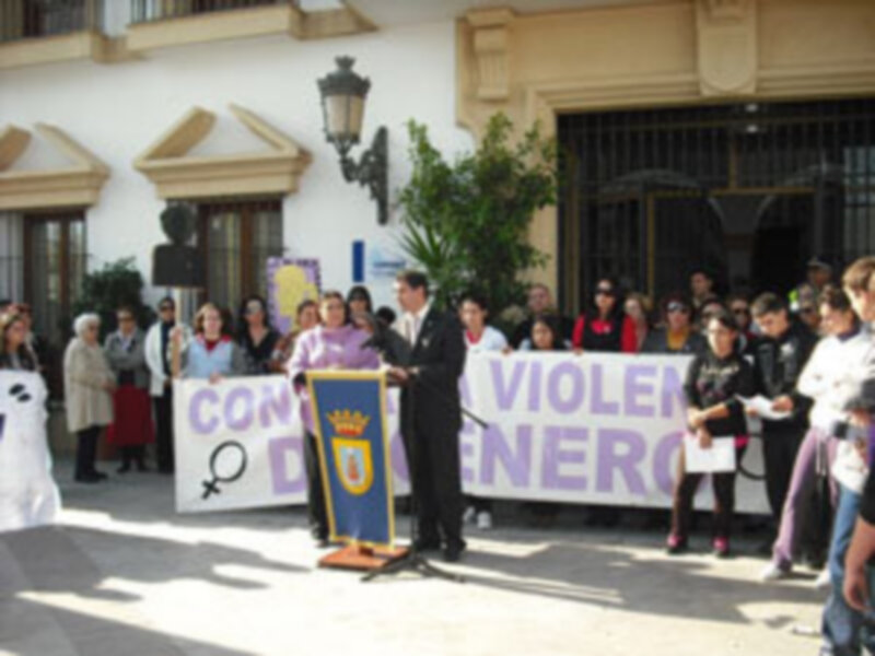 Una persona en un escenario sostiene un micrófono, frente a una pancarta que dice "Violencia de Género". Detrás hay personas con carteles y una bandera. La imagen parece ser de un evento o protesta contra la violencia hacia las mujeres.
