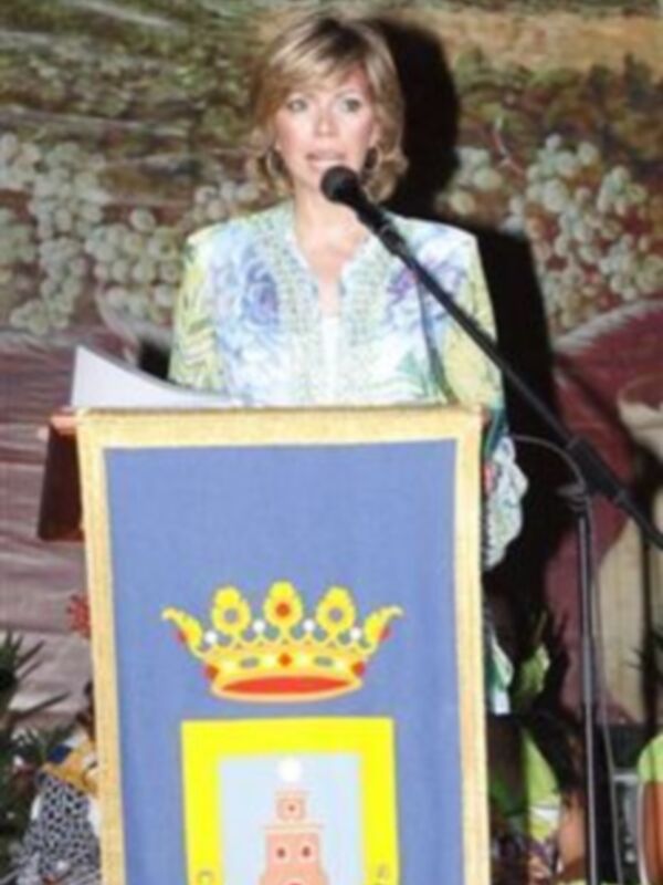 Una mujer habla en un evento formal, detrás de un escudo con una corona y un edificio, posiblemente un acto oficial o cerimonial.