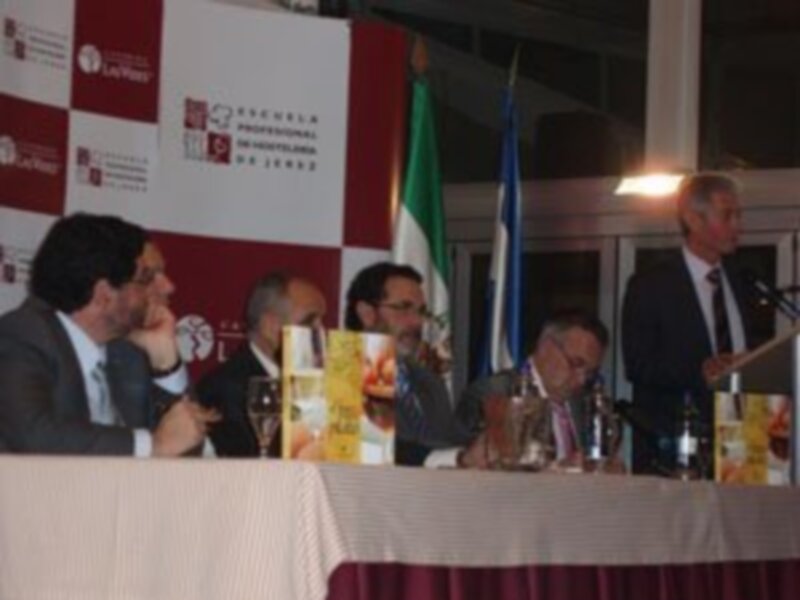 Conferencia o evento formal con banderas y logos en el fondo, participantes sentados en una mesa con documentos y vasos de vino.