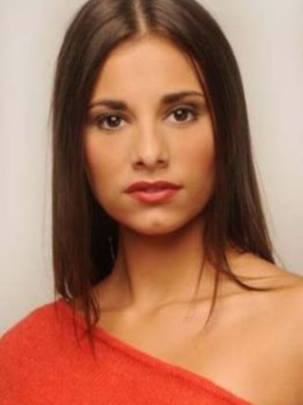 Una mujer con cabello largo y ondulado, vistiendo una blusa roja de hombros descubiertos.