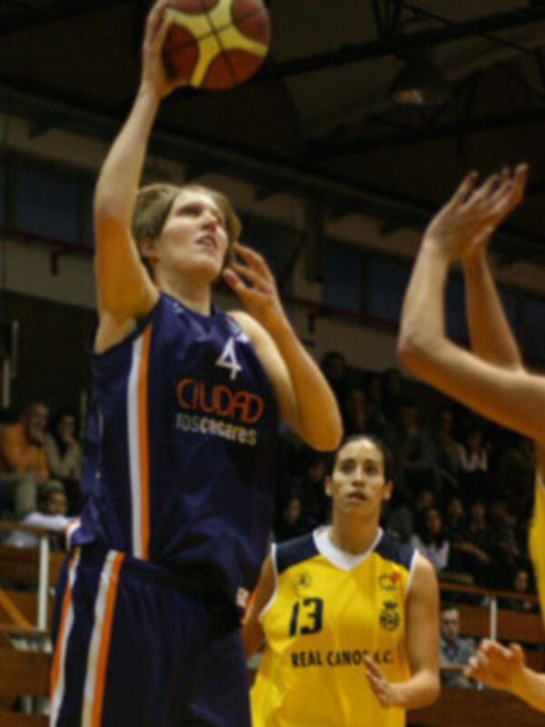 Jugador en acción durante un partido de baloncesto, lanzando al aro mientras un defensor intenta bloquear.