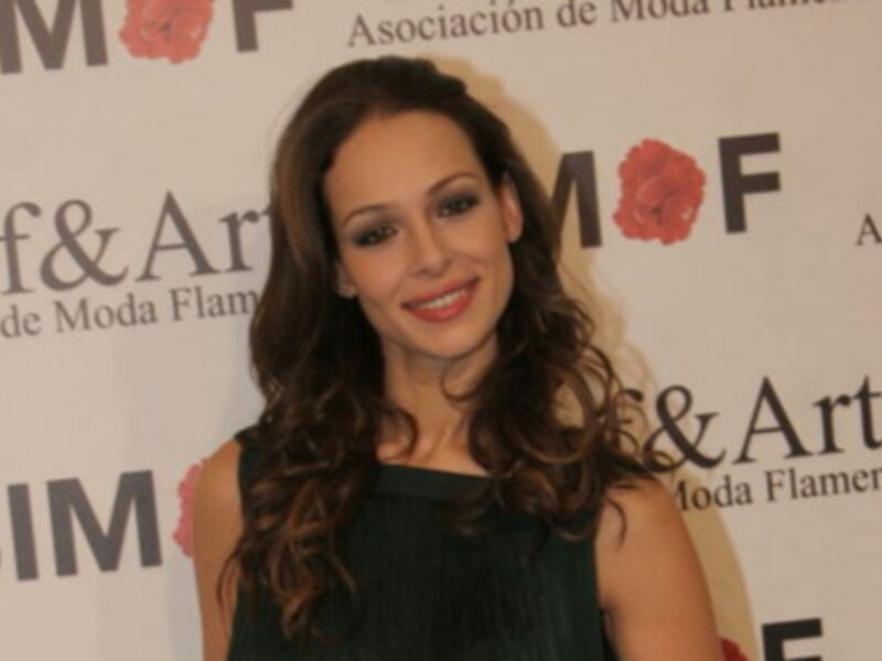 Una mujer con cabello largo y ondulado, maquillaje destacado y una sonrisa alegre frente a un fondo con el logo de la Asociación de Moda Flamenca.