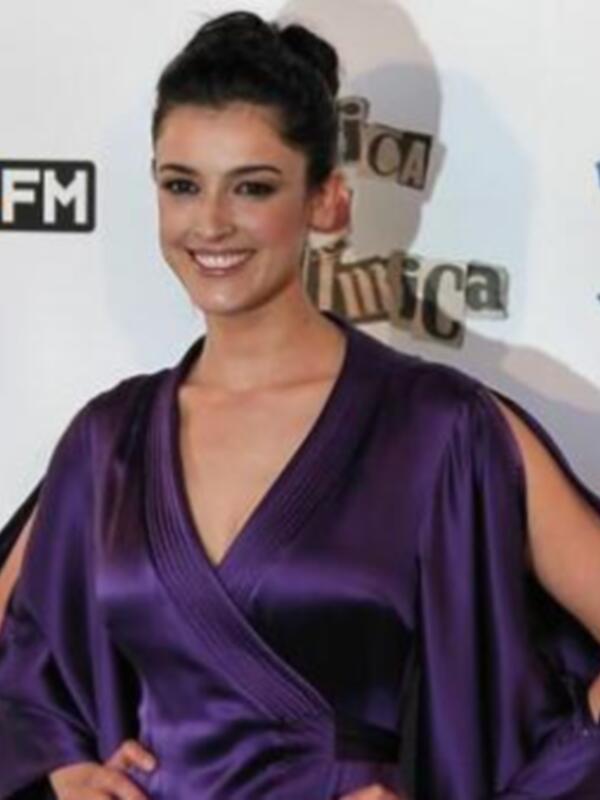 La modelo posa con un vestido púrpura en una alfombra roja, frente a un fondo con logos de radio y televisión.