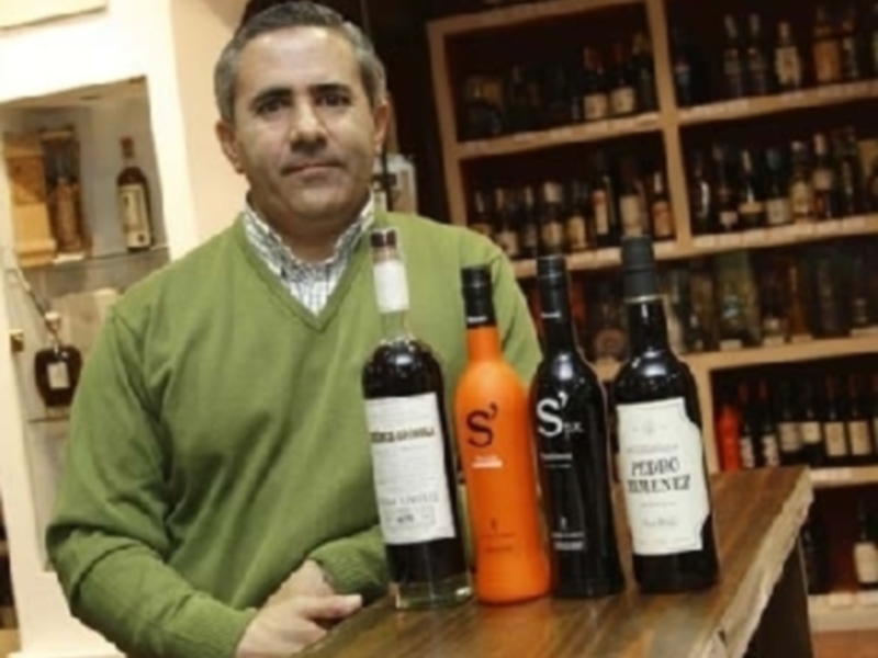 Un hombre sostiene botellas de vino en un entorno de bodega, rodeado de estantes de botellas.