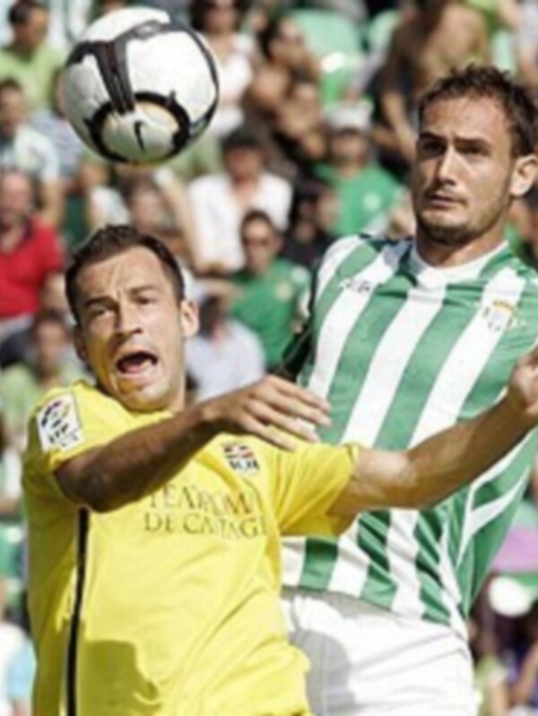 Jugadores en acción durante un partido de fútbol, uno en verde y blanco y otro en amarillo, disputando el balón en el aire.