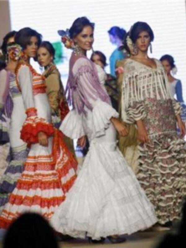 Una modelo camina por la pasarela en un desfile de moda, luciendo un vestido blanco con volantes y detalles en rojo y blanco. Detrás de ella, otras modelos también lucen vestidos de estilo clásico y colorido, con volantes y detalles en azul y blanco.