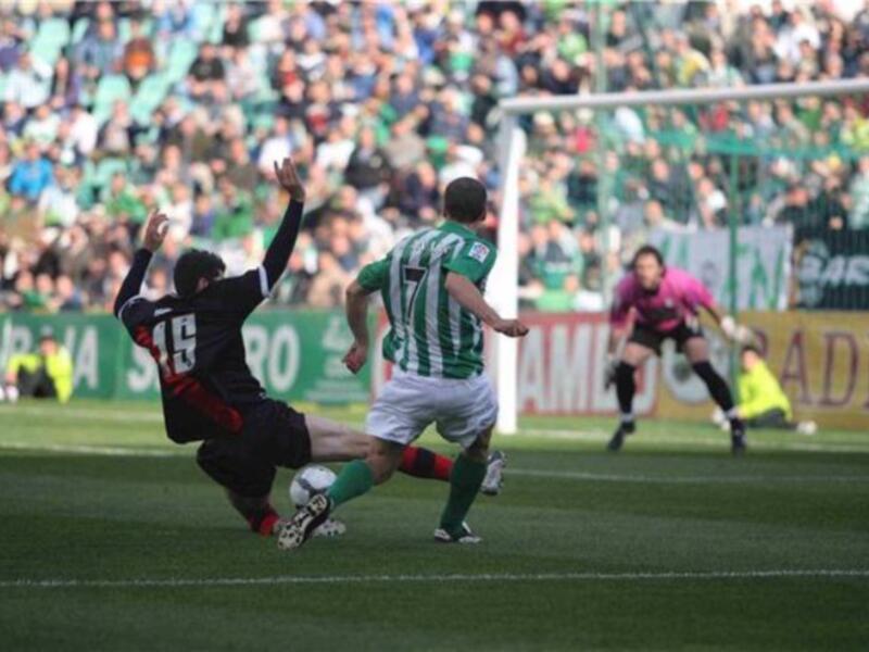 Un jugador en verde y blanco intenta controlar el balón durante un partido de fútbol mientras un rival en negro y rojo intenta despejarlo.