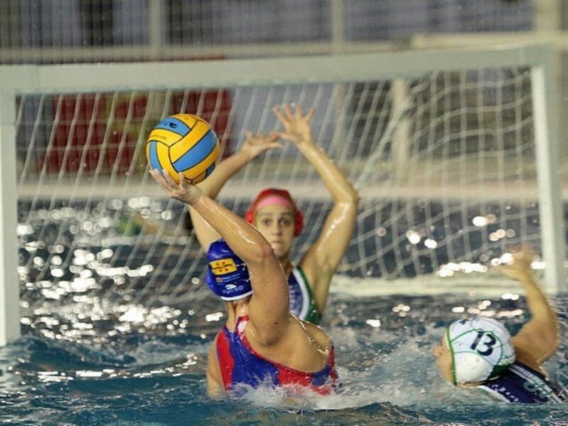 Jugadoras de waterpolo compiten por el balón en el agua, con un golero en el fondo.