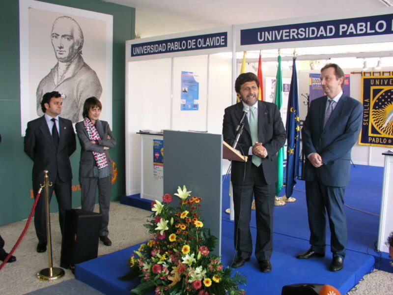 Presentación en la Universidad Pablo de Olavide, con un hombre en el escenario y dos personas de pie al lado, en un ambiente formal con banderas y flores en el centro.