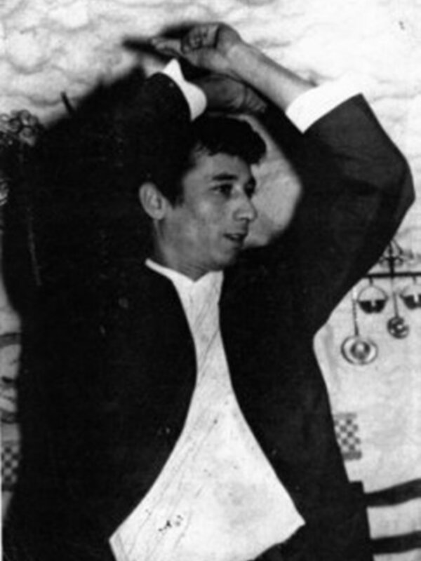 Un hombre con una camisa blanca y un traje negro, posando con las manos levantadas. El fondo parece ser una sala de baile o un salón decorado con elementos vintage.