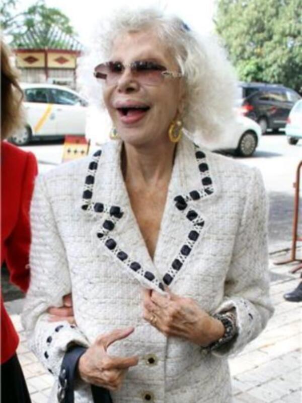 Una mujer con cabello blanco largo y gafas de sol, vistiendo un abrigo blanco con detalles negros.