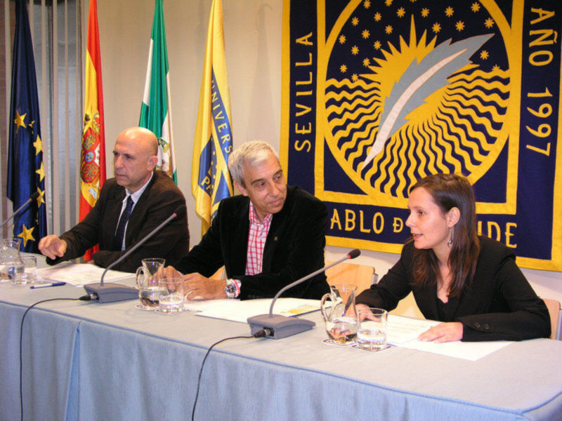 Tres personas sentadas en una mesa con banderas y un escudo de Sevilla como fondo, durante una reunión o conferencia.