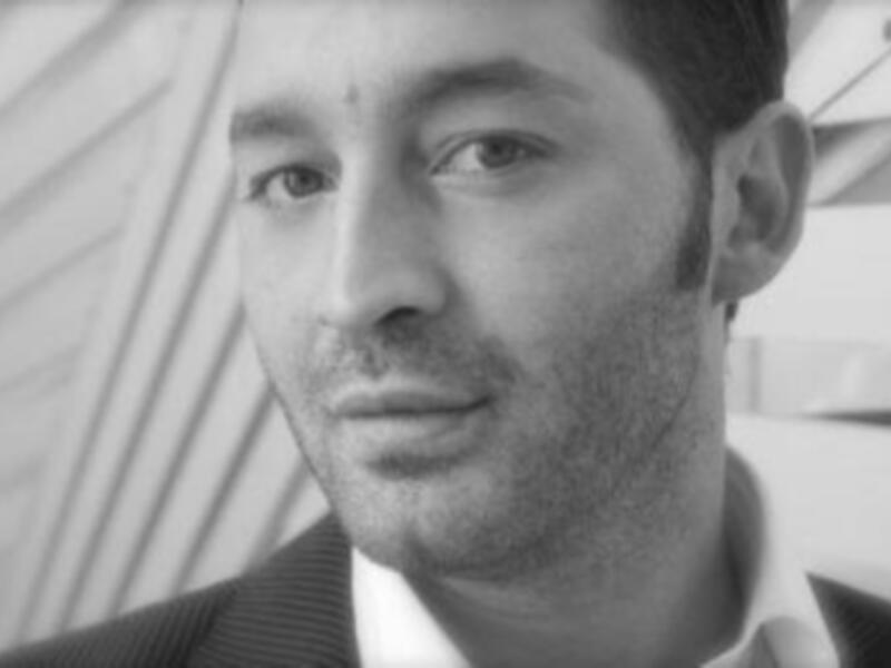 Retrato en blanco y negro de un hombre con barba, vistiendo traje formal, con fondo abstracto de líneas diagonales.