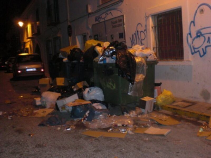 Basura acumulada en contenedores y suelo de una calle nocturna.