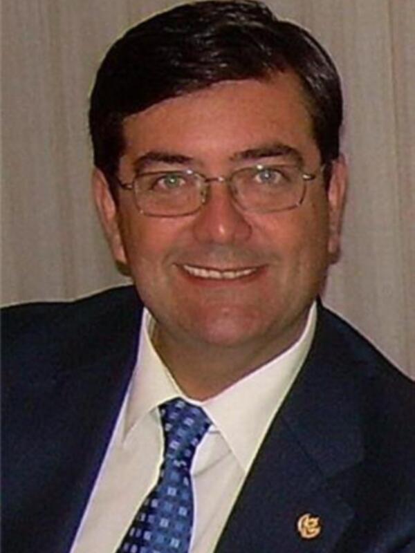 Retrato de hombre sonriente en traje y corbata, con logotipo en el pecho.