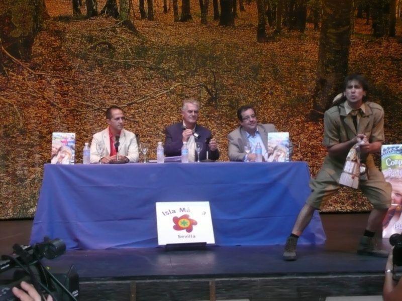 Una conferencia de prensa en un escenario con tres personas sentadas en una mesa y un hombre de pie a su lado, con un fondo de árboles y hojas caídas en el suelo.
