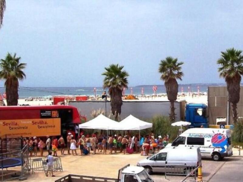 Playa de Sevilla con palmeras, una camioneta y un globo en el cielo, con la playa y el mar al fondo.