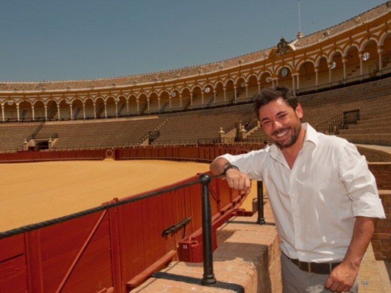 Un hombre en una camisa blanca y pantalones beige se encuentra en el interior de un estadio de arena, posando con una sonrisa. La arena es de color amarillo y el estadio tiene arcos dorados en la parte superior.