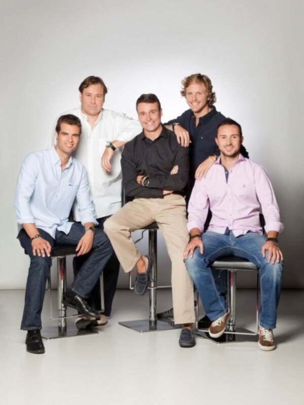 Cuatro hombres sonrientes sentados en taburetes, vestidos con camisas y pantalones de estilo casual, en un fondo blanco.