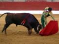 Un toro negro con una flecha roja en el pecho se enfrenta a un matador de toros vestido con traje dorado y verde, que sostiene una muleta roja. La escena se desarrolla en un anfiteatro de arena con una pared roja al fondo.