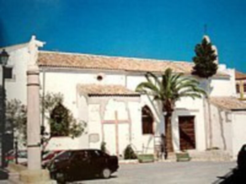 Iglesia antigua con fachada blanca y cruz en el techo, rodeada de palmeras y un vehículo en la entrada.