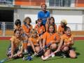 Una escuela de hockey infantil posa en un estadio deportivo, con niños y niñas vestidos con uniformes naranjas y azules. Al fondo, un hombre en camiseta azul supervisa la escuela.