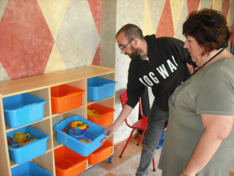 Un hombre y una mujer están jugando con un cubo de arena en un estante de almacenamiento lleno de cajas de colores. La pared tiene una textura geométrica en tonos pastel.