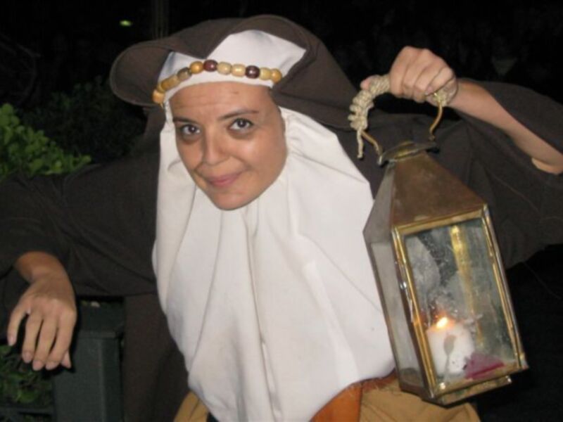 Una persona vestida con un traje medieval, sosteniendo una linterna con un conejo dentro, en una noche oscura con plantas verdes al fondo.